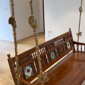 Traditional wooden swing (jhoola) with brass chains and hand-painted tile inlays, suspended in a modern indoor space.