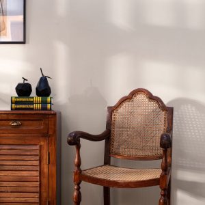 A classic wooden armchair with cane weaving, placed beside a wooden side cabinet topped with decor items and books, against a light-colored wall.