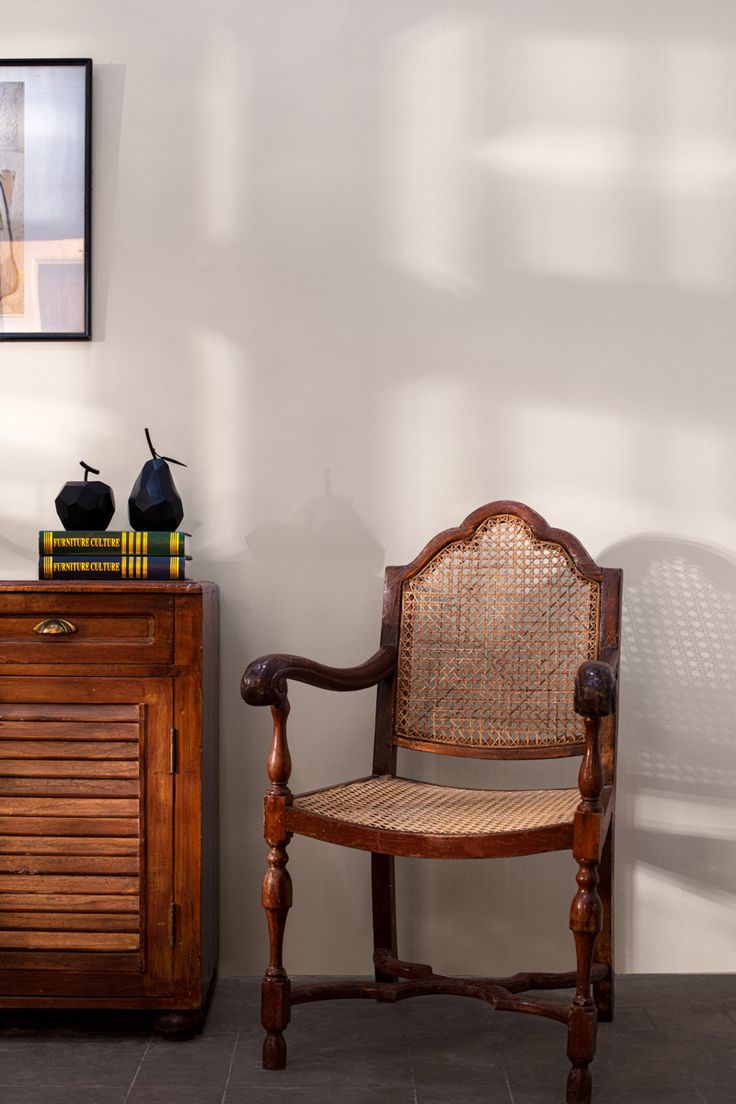 Modern Indian Home _ Dining A classic wooden armchair with cane weaving, placed beside a wooden side cabinet topped with decor items and books, against a light-colored wall.